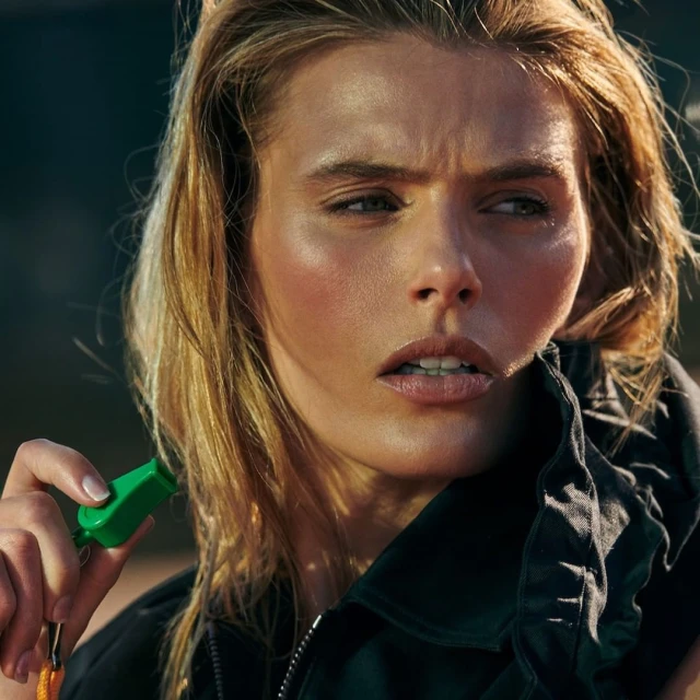 Blonde-haired adult woman in a jacket holds a green whistle with orange lanyard against an outdoor background in a medium shot.