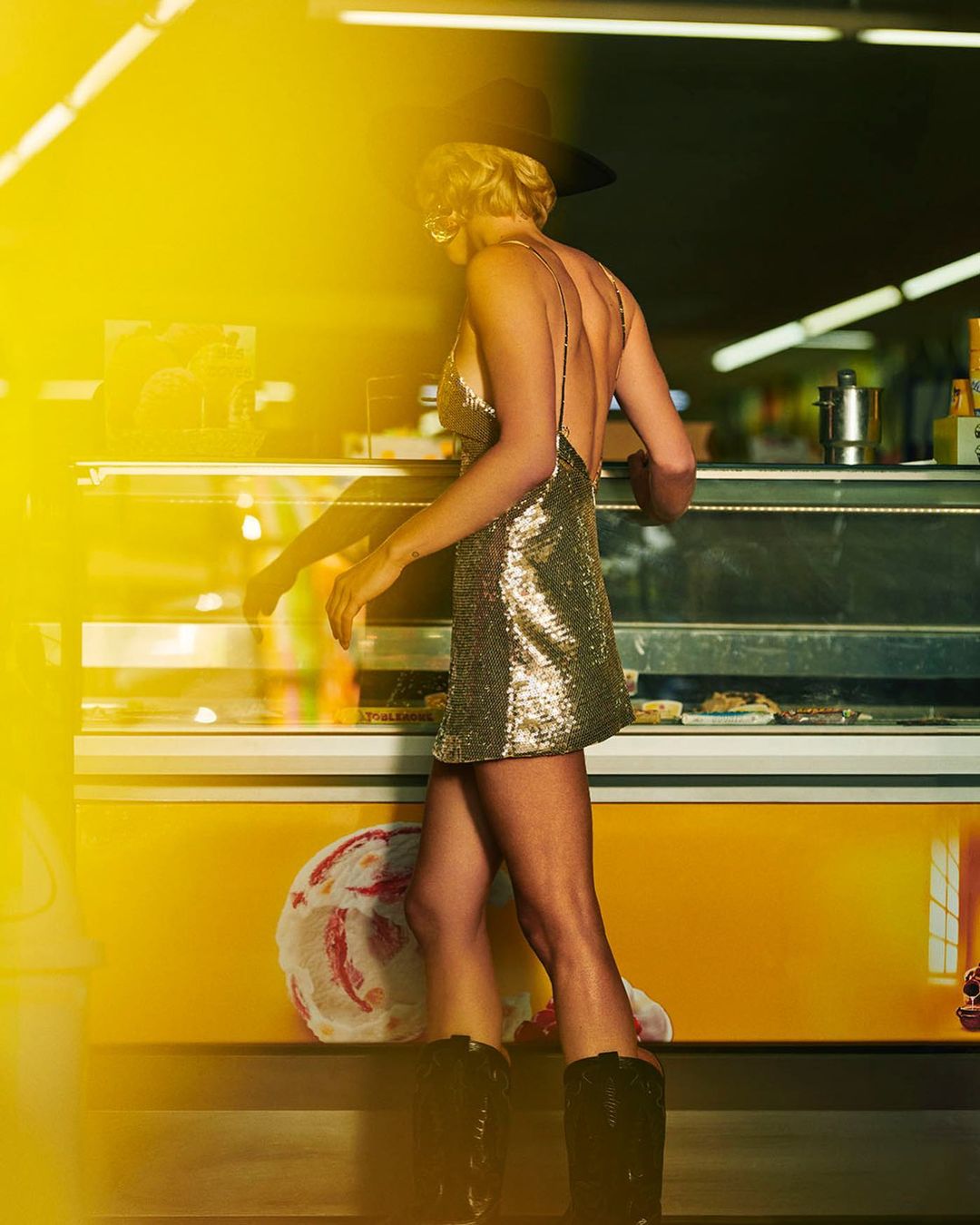 Woman in a gold dress and black boots stands near a yellow ice cream cooler under bright lighting, seen from the back.
