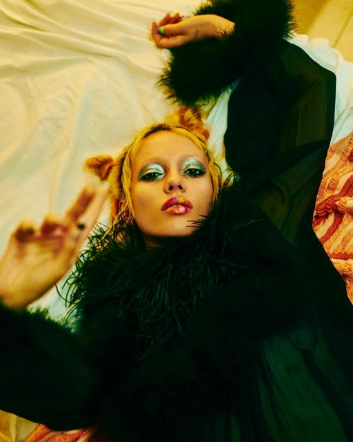 Woman with creative eye makeup and a black feathered top laying on a bed with a sheet and a striped towel, holding up a peace sign.