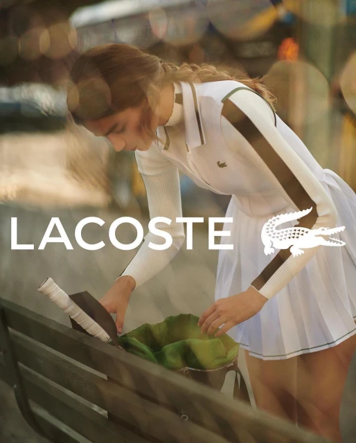 A fashionable young woman in a white tennis dress and Lacoste branding, reaching into a tennis bag, with an outdoor backdrop and soft focus.
