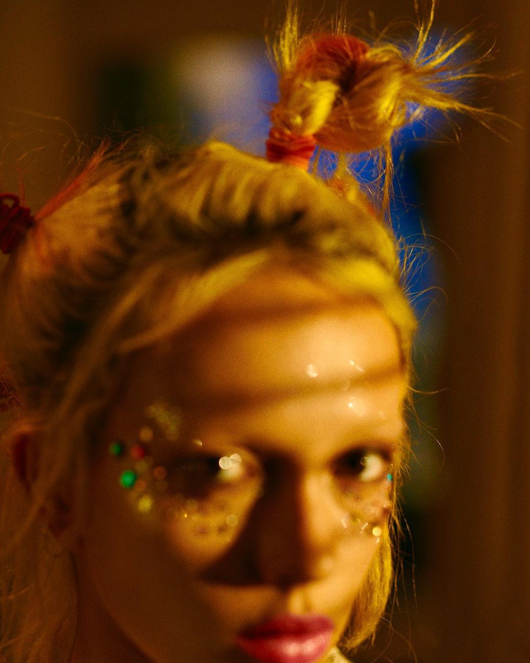 Close-up portrait of a blonde adult female with glitter makeup around her eyes and a bright, warm lighting in an indoor environment.