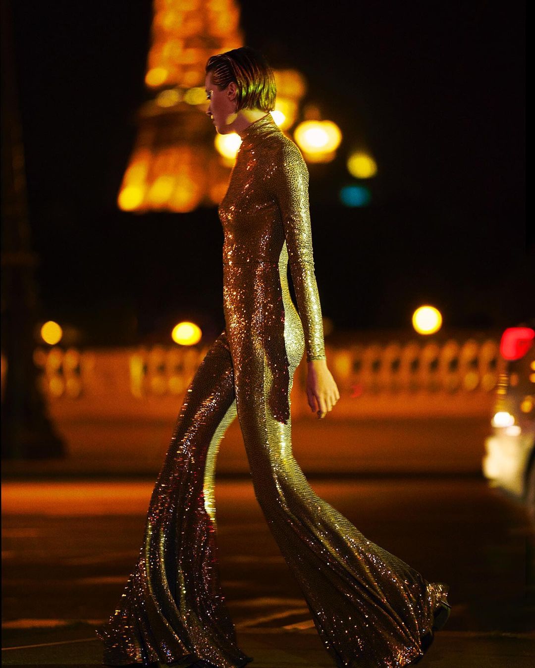 A woman in a gold sequined jumpsuit walks at night with the Eiffel Tower illuminated in the background, showcasing fashion in Paris.