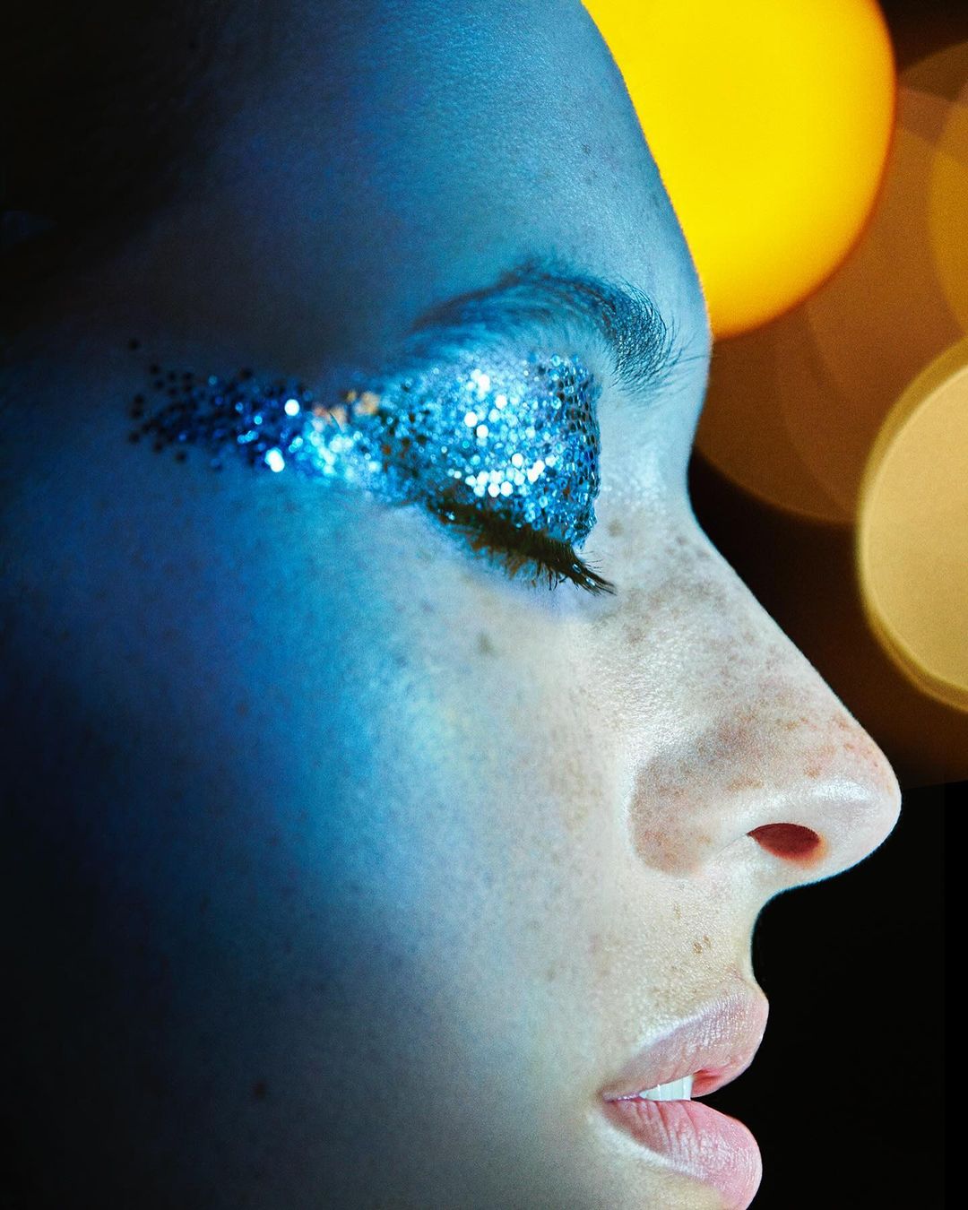 Close-up portrait of a young adult woman with glittery blue eye makeup, closed eyes, shot in a photo studio with a dark background.