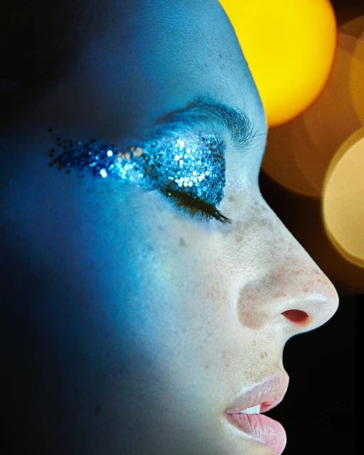 Close-up portrait of a young adult woman with glittery blue eye makeup, closed eyes, shot in a photo studio with a dark background.