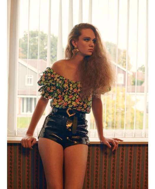 Woman with curly hair poses in floral blouse and black sequin shorts with heart belt indoors, in front of a window with sheer curtains.