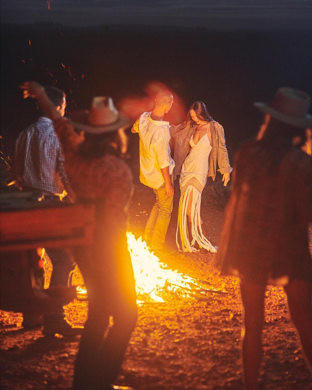 A group of people gather around a bonfire in a rural setting at night, illuminated by the warm glow of the flames and dark skies.