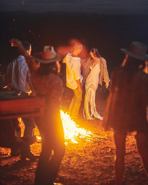 A group of people gather around a bonfire in a rural setting at night, illuminated by the warm glow of the flames and dark skies.