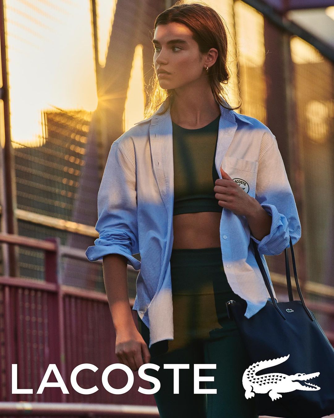 Athletic woman in workout clothes and unbuttoned shirt carrying a tote bag, captured in an urban setting with golden sunlight.