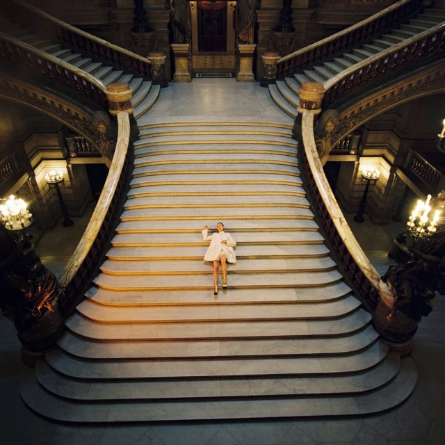 Woman in a white coat ascends a grand marble staircase, adorned with ornate gold details and chandelier lighting in a historical building.