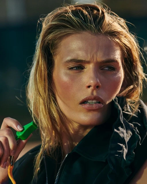 Close-up portrait of a blond adult female holding a green whistle attached to a yellow lanyard, wearing a dark jacket.