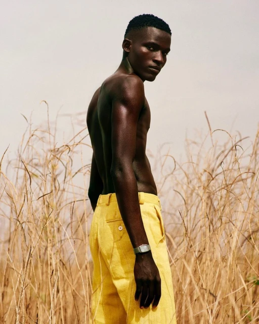 Portrait of a shirtless African male model wearing yellow pants, standing outdoors in a field with tall brown grass, looking at the camera.