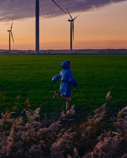 A child in a blue dress runs through a green field at sunset, with wind turbines in the background. A sense of freedom and energy.