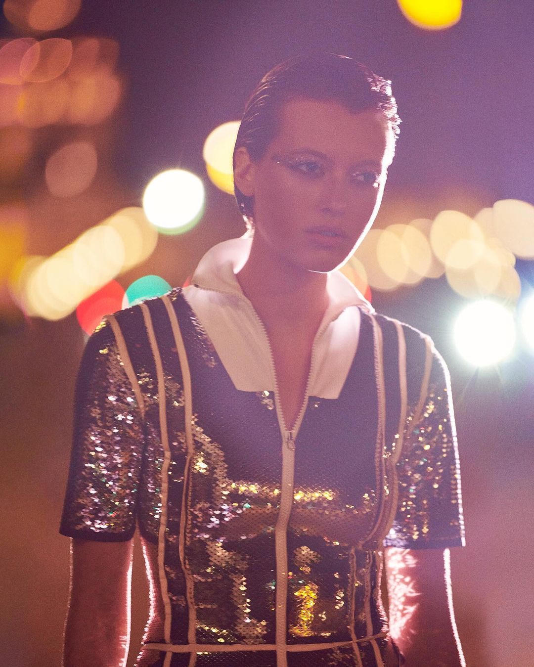 Close-up of a fashion model in a sparkly top with a white collar, posing outdoors at night with blurred lights in the background.