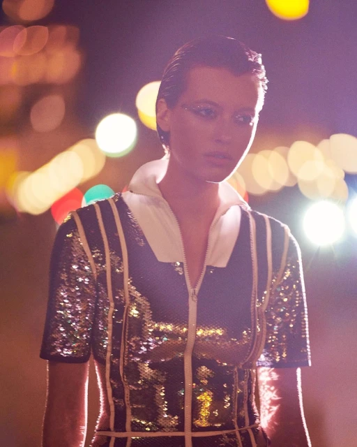 Close-up of a fashion model in a sparkly top with a white collar, posing outdoors at night with blurred lights in the background.