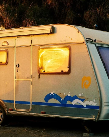 Side view of a vintage caravan with a blue and yellow wave design, parked outdoors with sunlight reflecting in the window, set against trees.