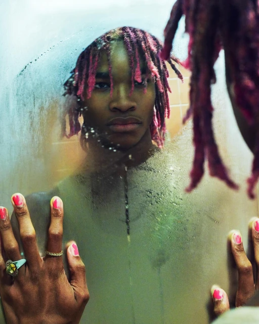A young adult stares with pink dreadlocks in an indoor portrait. The reflection in a wet mirror shows the subject and ornate rings.