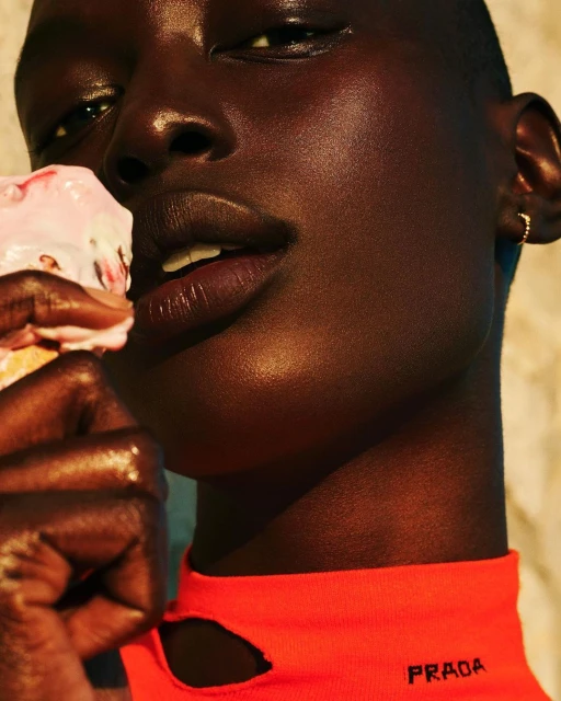 Close-up of a dark-skinned woman holding a melting ice cream cone, wearing a bright-orange top with the PRADA logo on it.