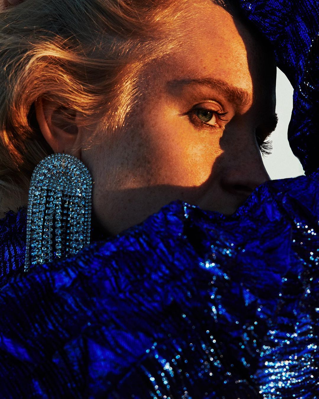 Close-up portrait of a blond woman with an earring, wearing a blue sequined garment, illuminated by warm, diffused lighting.