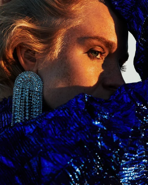 Close-up portrait of a blond woman with an earring, wearing a blue sequined garment, illuminated by warm, diffused lighting.