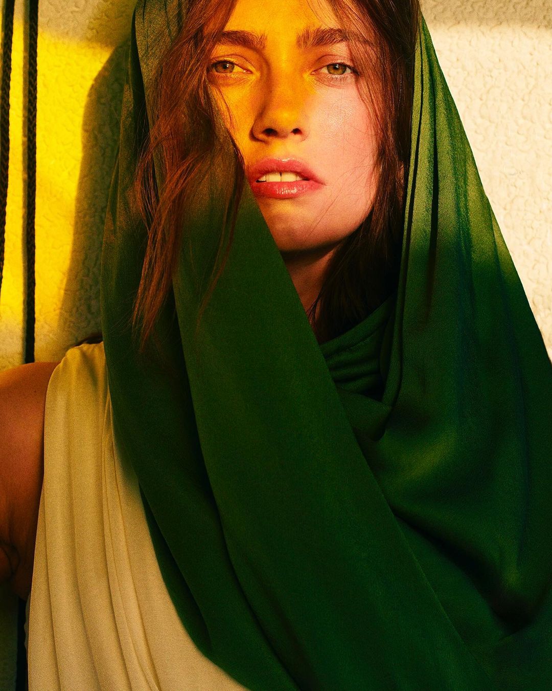 Woman with brunette hair and green headscarf gazes directly at the camera in a close-up shot with split yellow and green lighting.