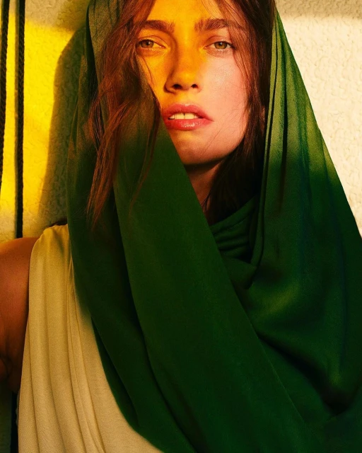 Woman with brunette hair and green headscarf gazes directly at the camera in a close-up shot with split yellow and green lighting.