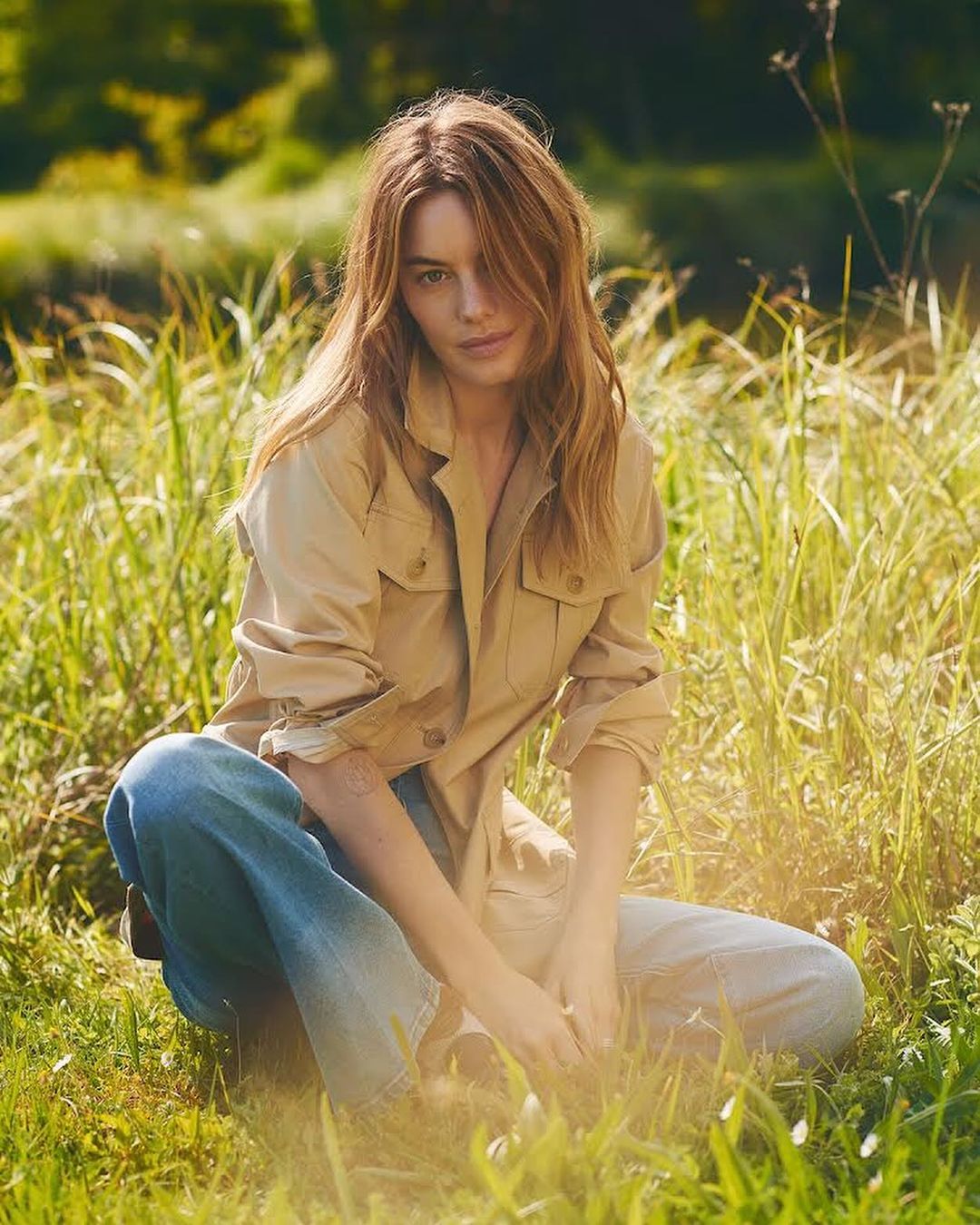 Woman with long brown hair wearing a tan utility jacket and blue denim jeans crouching in tall green meadow grass.
