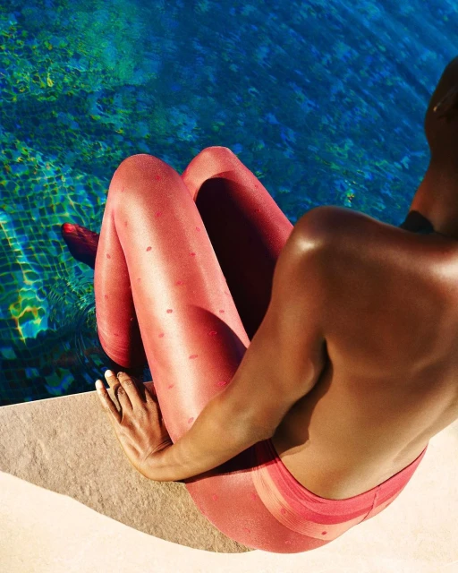 Woman in red patterned tights sits poolside on a sunny day, turquoise water glistening in the background, creating a vibrant summer scene.