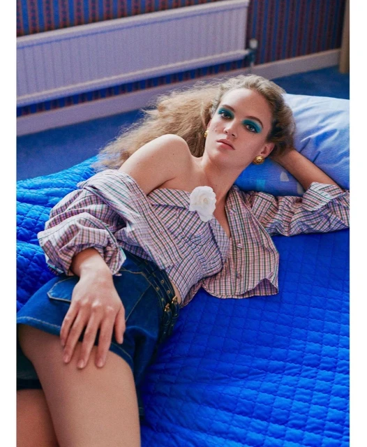 Woman lying on a bed wearing a plaid shirt and denim shorts, looking at the camera with soft blue and green eyeshadow makeup.
