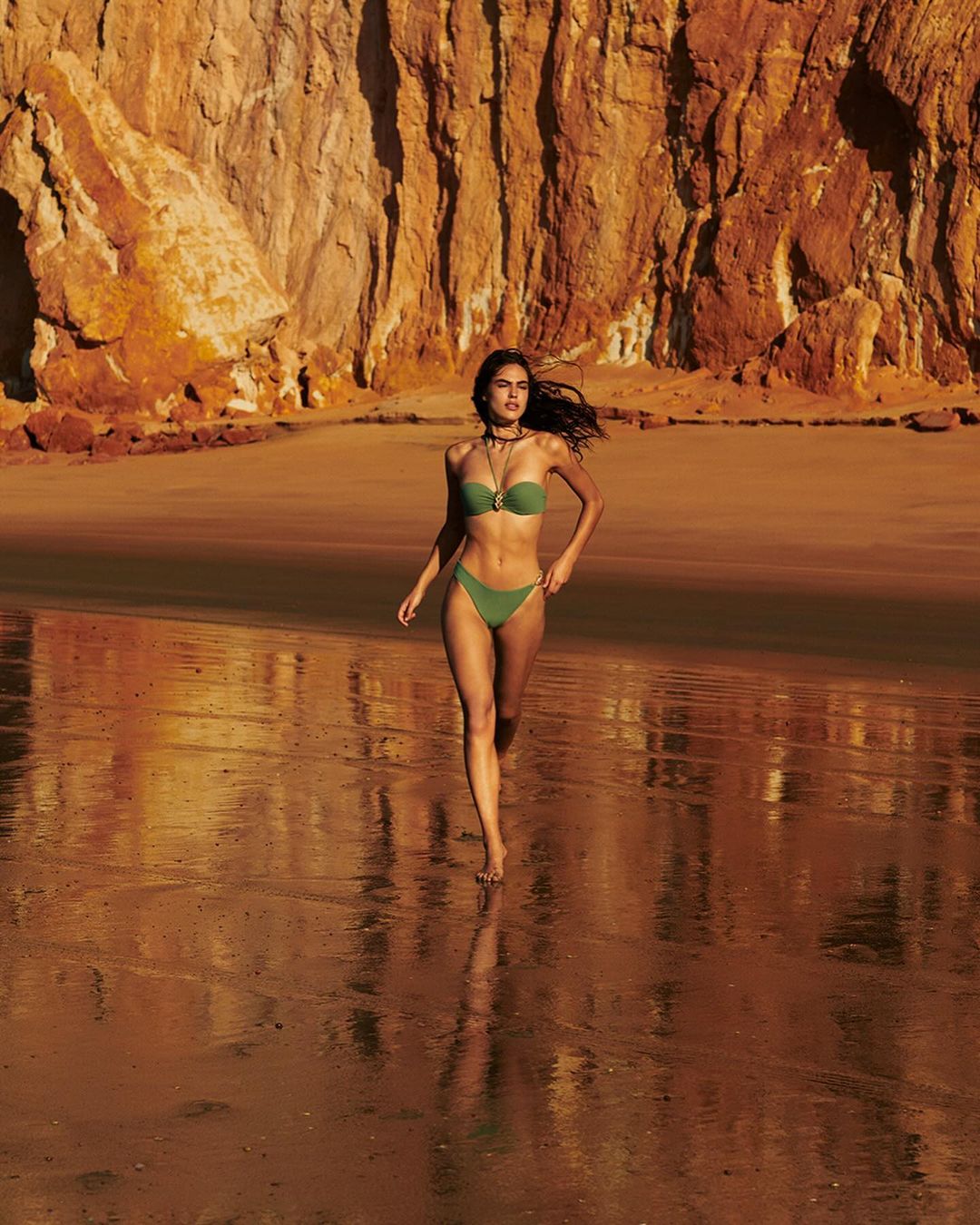Woman in a green bikini walking barefoot on a wet beach with a large red rock cliff in the background under bright sunlight.