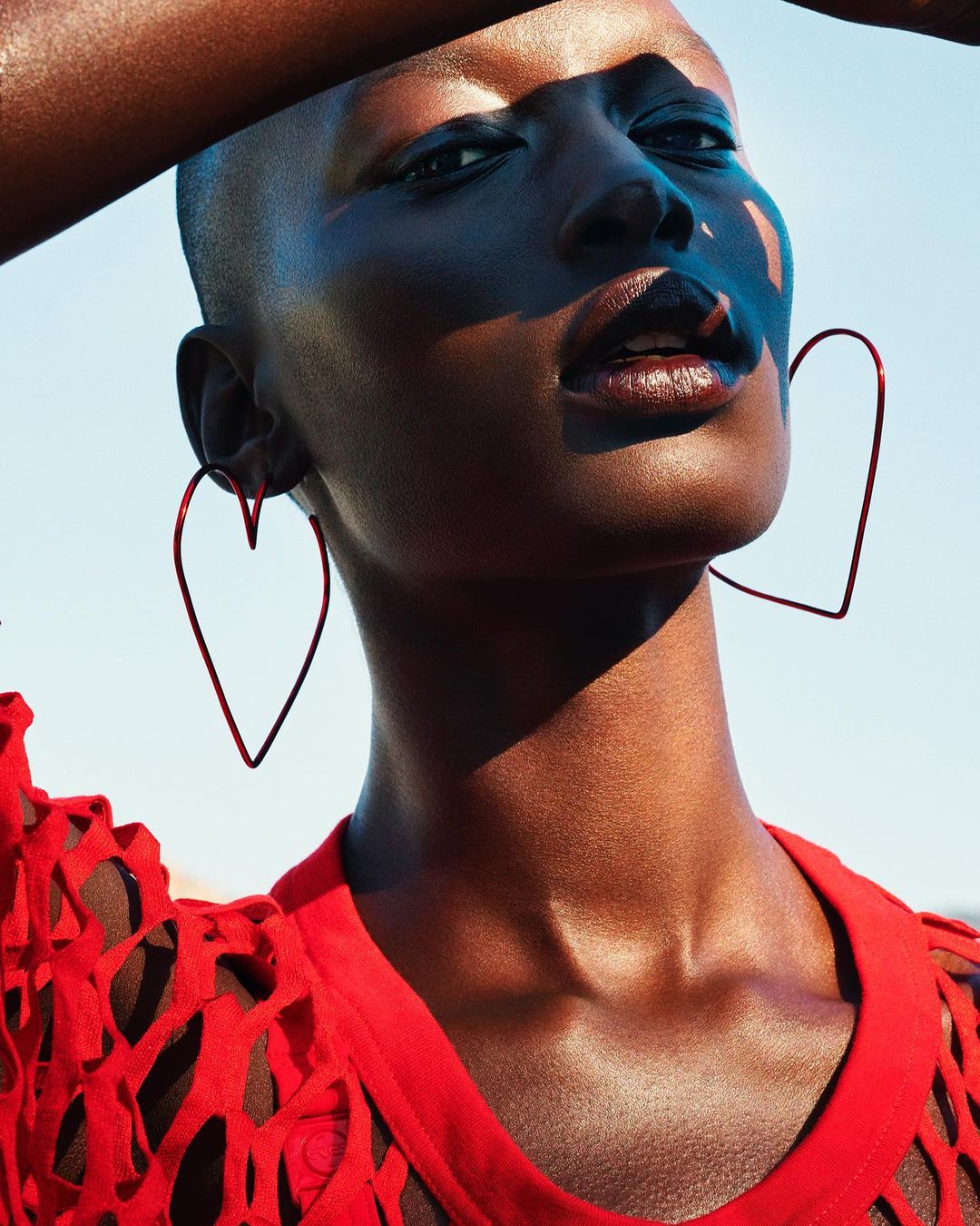 Fashion portrait of an African woman with short hair wearing heart shaped earrings and a red top against a blue sky backdrop.