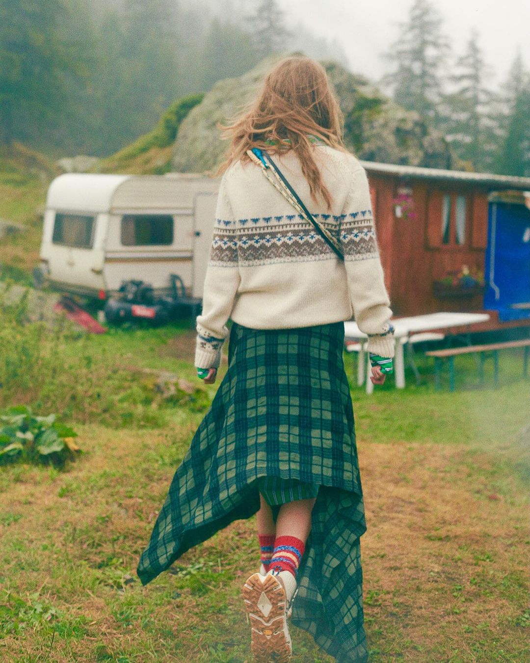A woman with long hair walks through a grassy field. She wears a patterned sweater and plaid skirt, with a caravan and trees behind her.