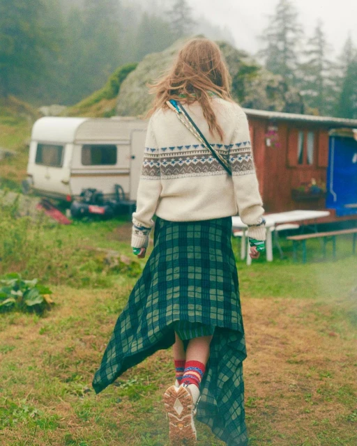 A woman with long hair walks through a grassy field. She wears a patterned sweater and plaid skirt, with a caravan and trees behind her.