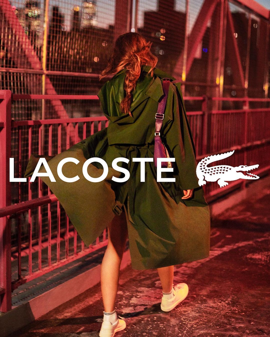 A woman walks on a bridge wearing a green coat and white sneakers, featuring the Lacoste logo and brand name in an outdoor setting.