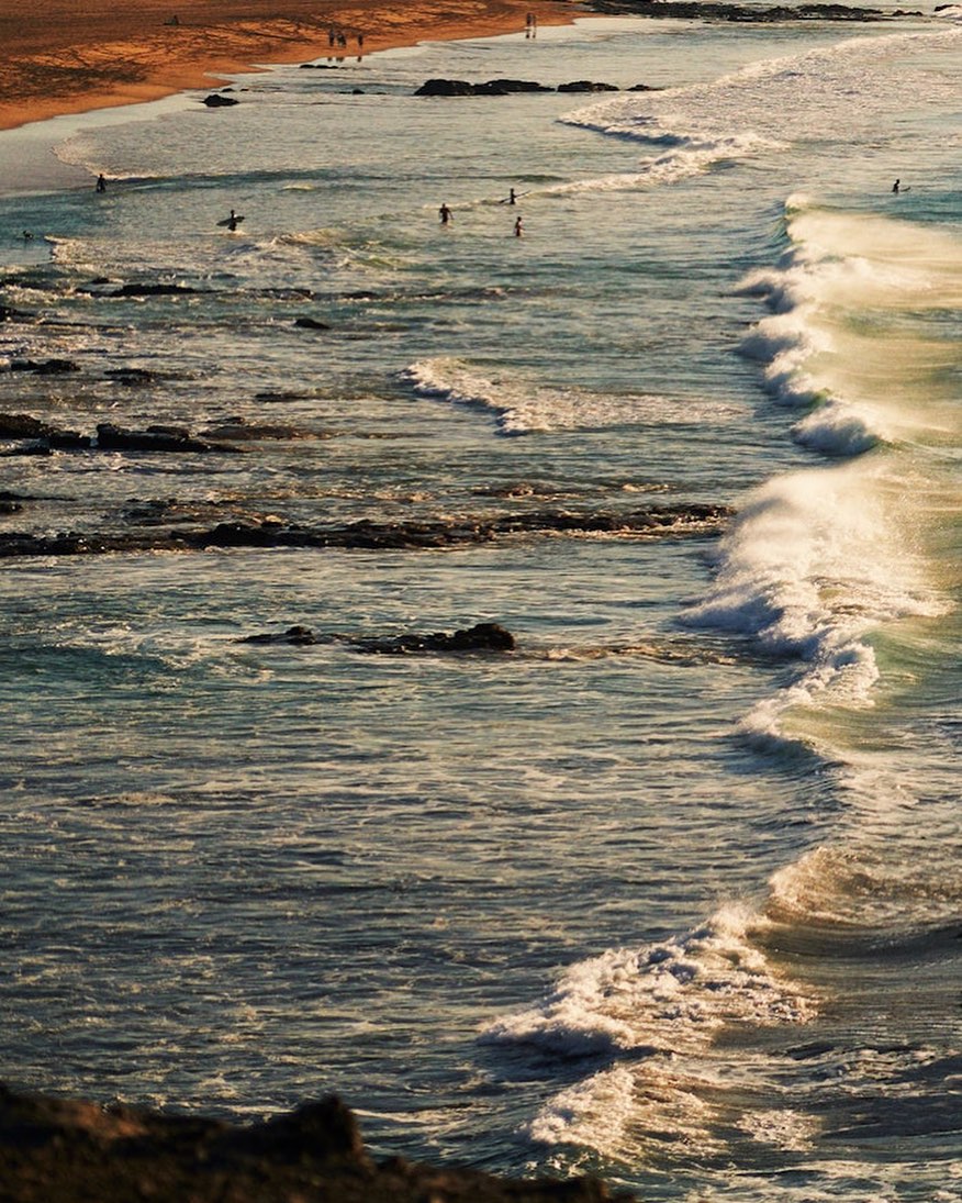 Seascape photo of crashing waves on a sandy shore with people in the distance, captured at eye-level in natural light.