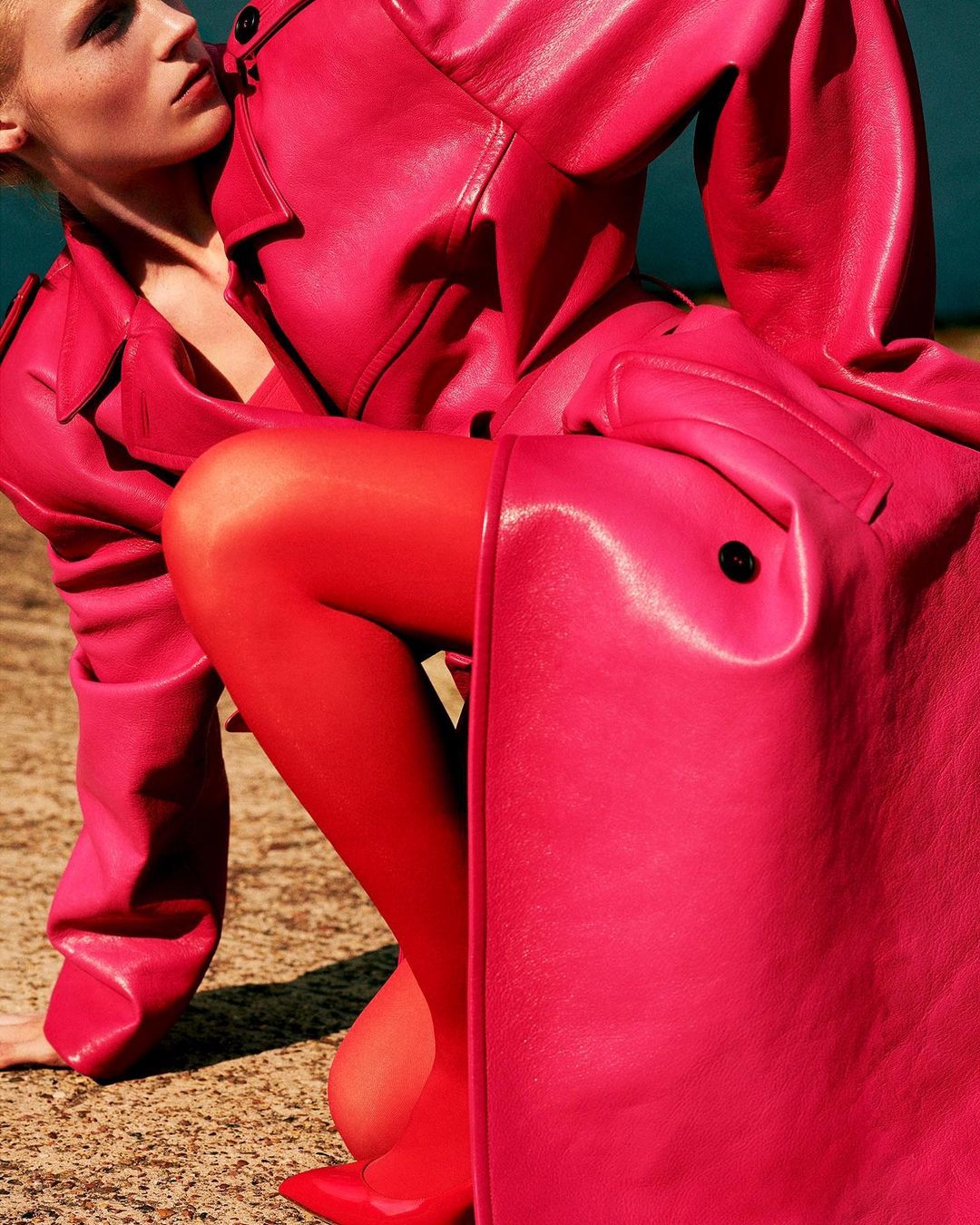 Two women pose wearing matching bright pink leather trench coats and red stockings in an outdoor setting with a neutral background.