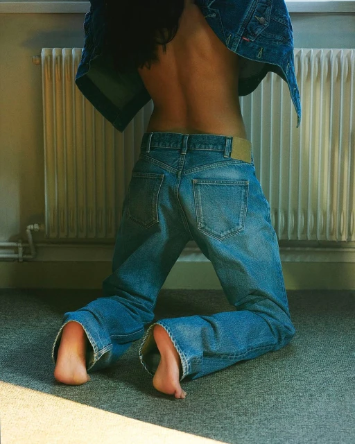 Young woman kneeling indoors, wearing denim jacket and jeans, showcasing bare back and feet, lit by natural light.