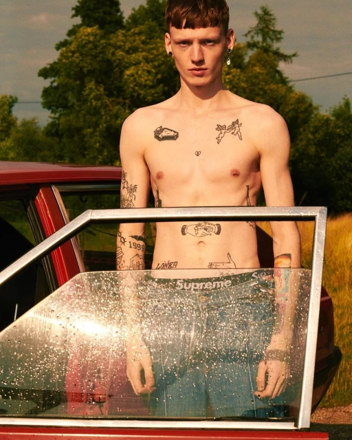 Shirtless man with tattoos poses near a red car in an outdoor setting, showcasing fashion and style in a lifestyle image.