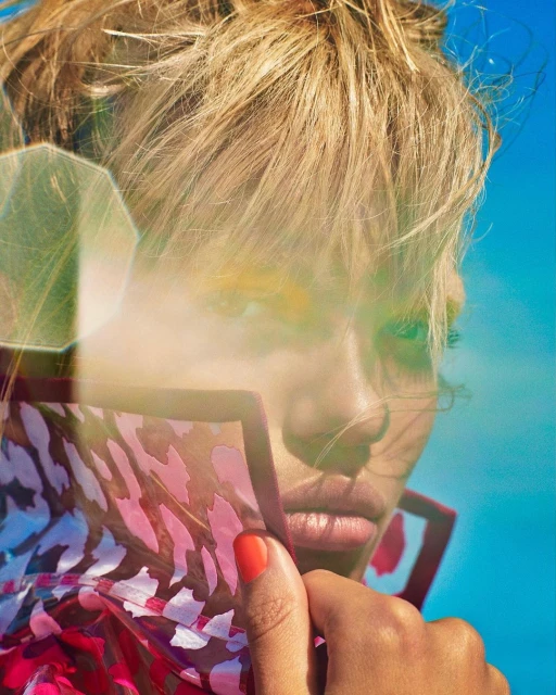 Close-up portrait of a blonde woman with a sheer patterned shawl, yellow eyeshadow, and red manicure, set against a vibrant blue sky.