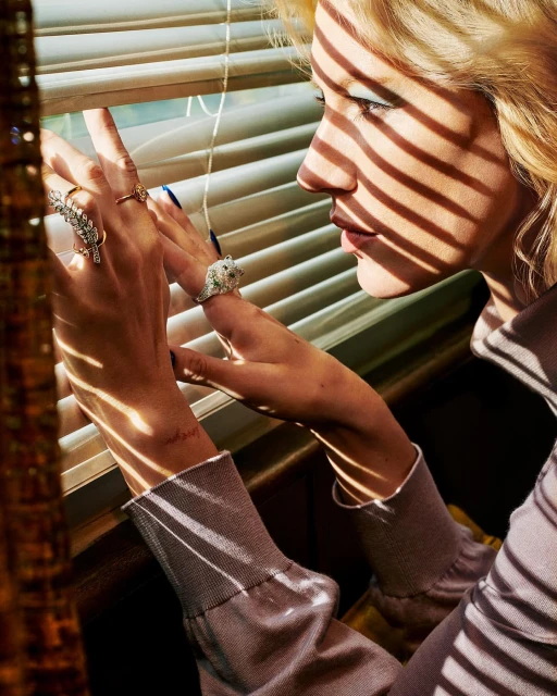 Close-up of a blonde woman's face and hands adorned with rings, looking out through blinds with sunlight creating shadows.