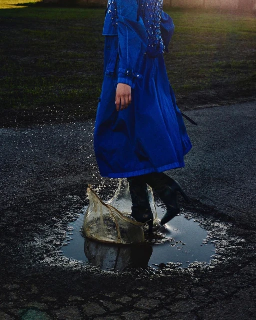 Adult female in blue coat jumps in a puddle on a dark asphalt surface, creating splash of water in outdoor setting with dark background.