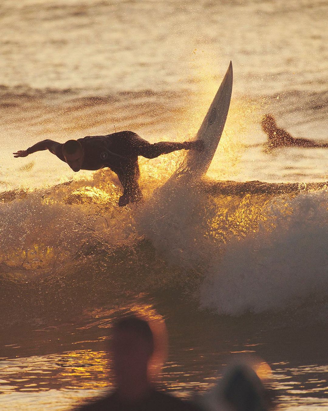 Surfer performs an aerial trick on a surfboard in golden light, with ocean waves and other surfers visible in the background.