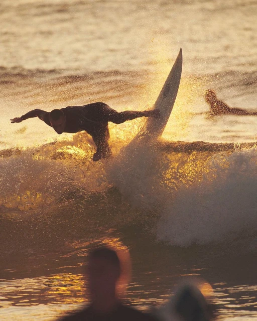 Surfer performs an aerial trick on a surfboard in golden light, with ocean waves and other surfers visible in the background.