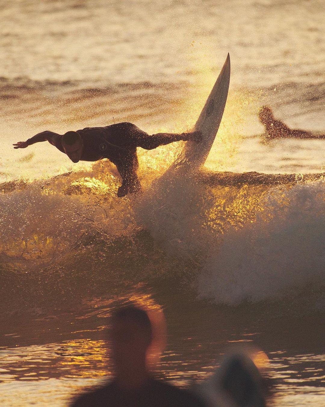 A surfer in a black wetsuit balances on a surfboard against the golden light of the ocean at sunset, with other surfers in the background.