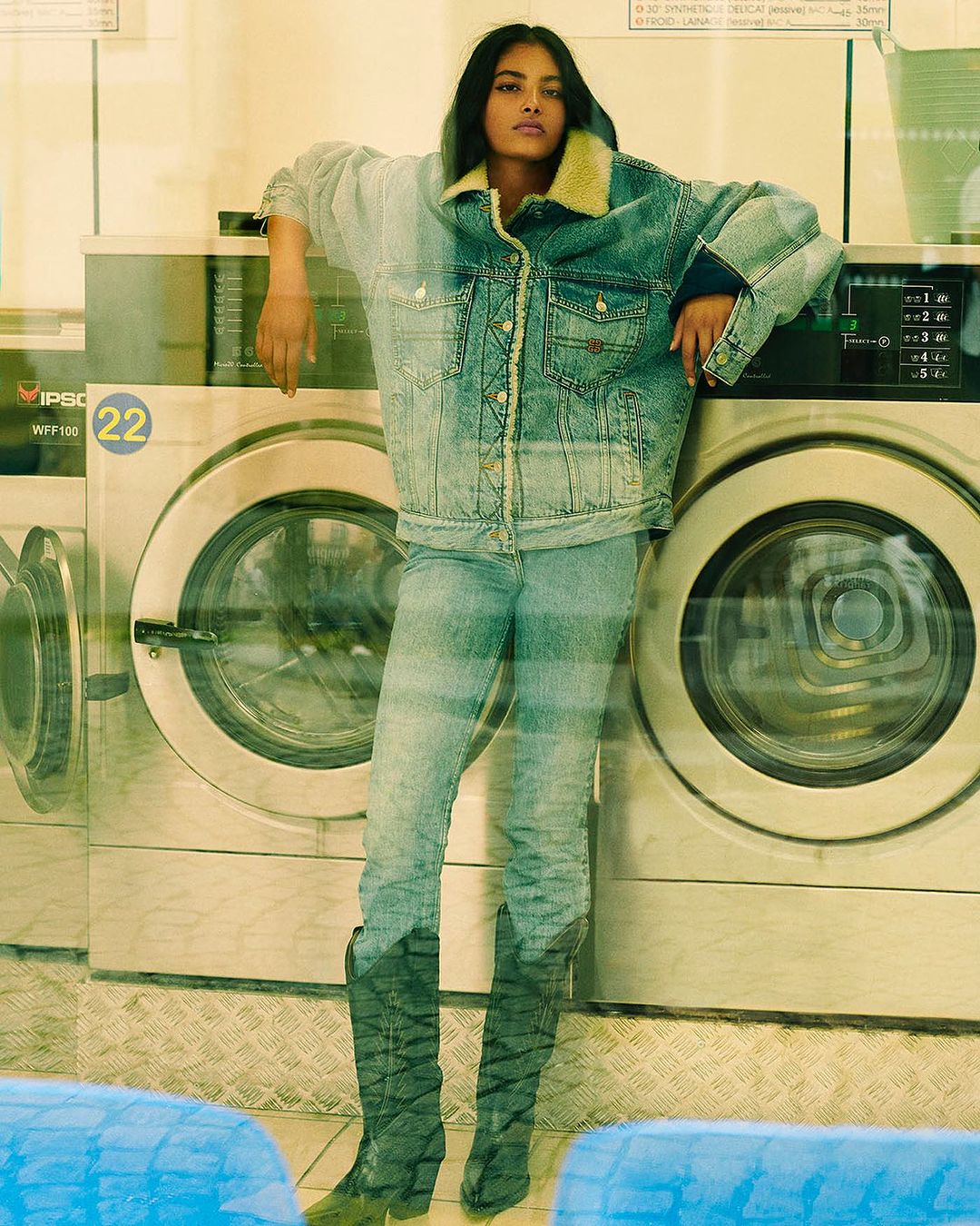Adult female fashion model in a denim jacket and jeans poses indoors against washing machines in a laundromat setting in daylight.