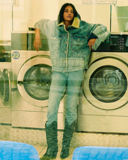 Adult female fashion model in a denim jacket and jeans poses indoors against washing machines in a laundromat setting in daylight.