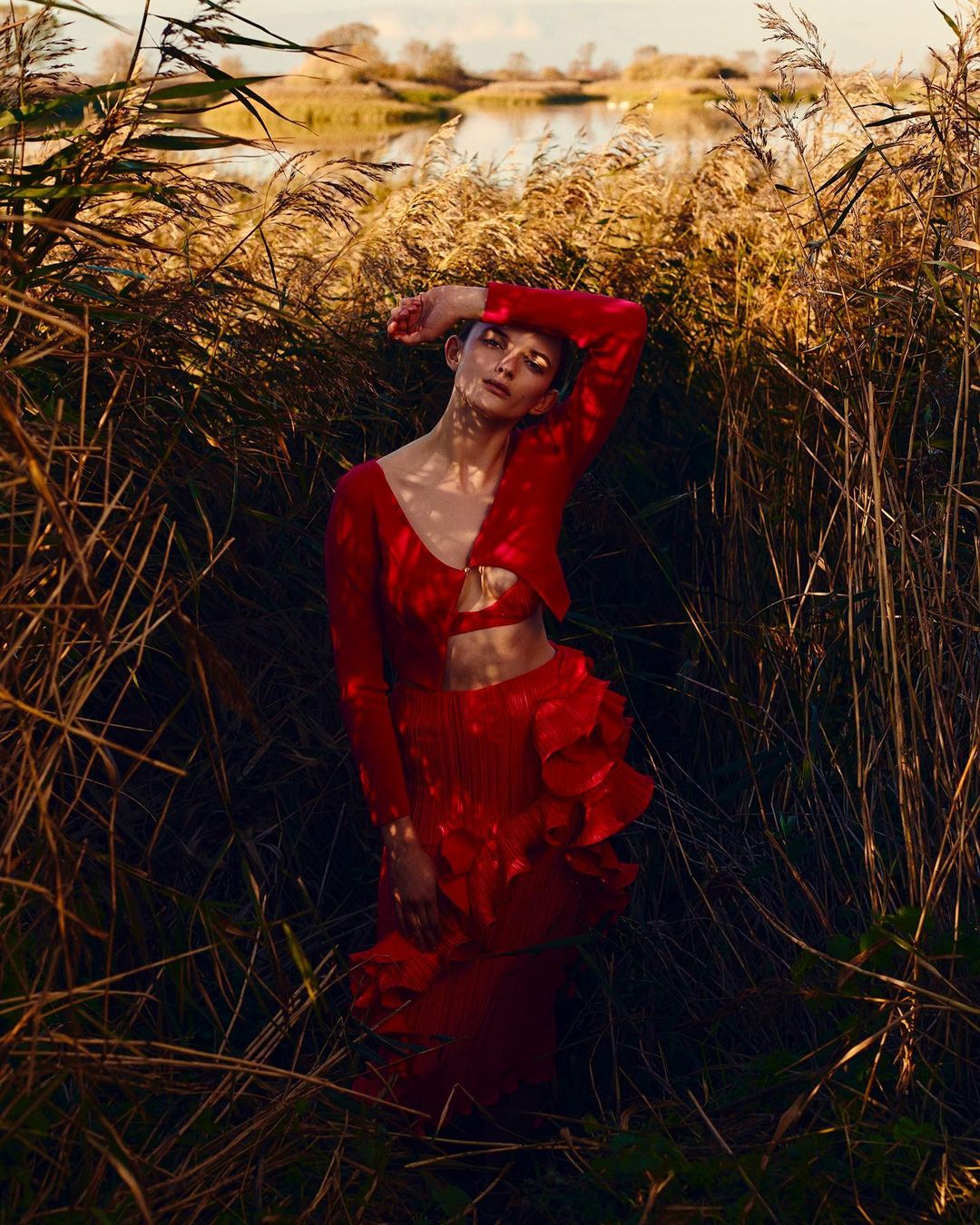 A young woman with her hand near her face is standing in tall grasses with a red dress and jacket and a lake in the background.