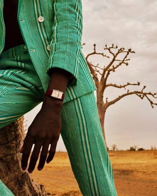 Person in a green striped suit and watch sits on tree stump in front of savannah landscape on a cloudy day in a fashion editorial.