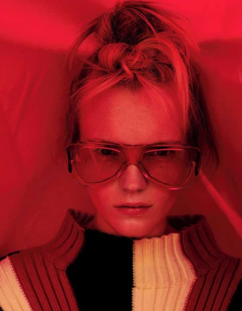 Portrait of a young adult woman with blonde hair in a high bun wearing large glasses and a black and white sweater against a red backdrop.