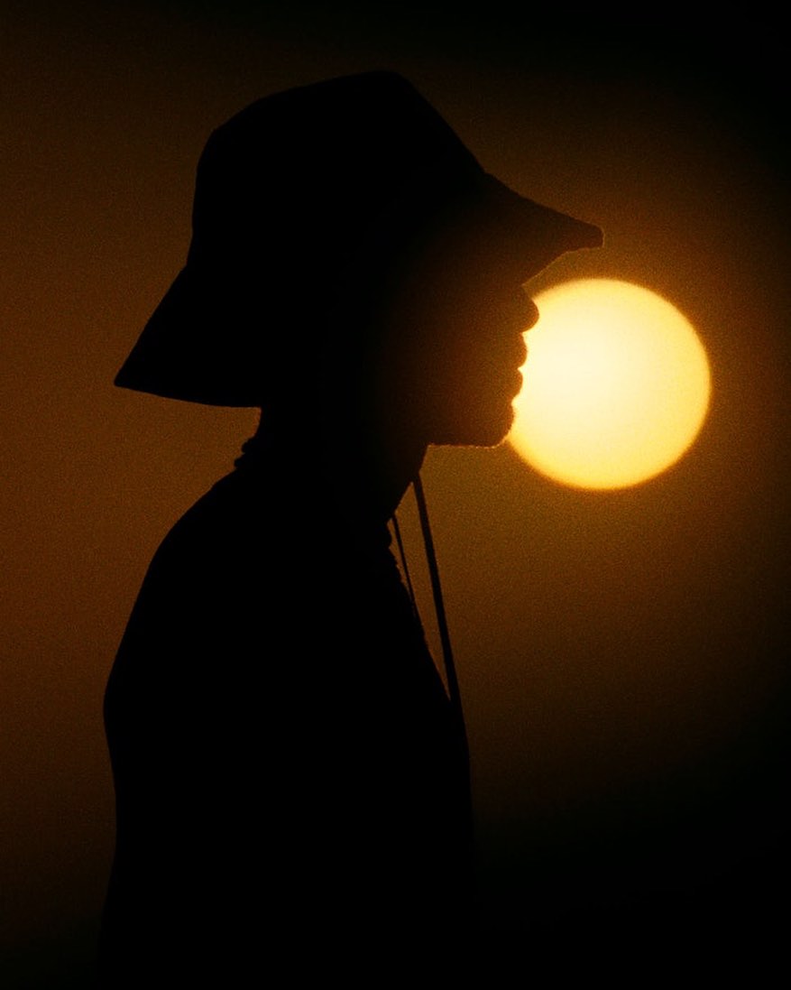 Silhouette of a person wearing a hat against a golden sky with the sun in the background, creating a warm and hazy atmosphere.