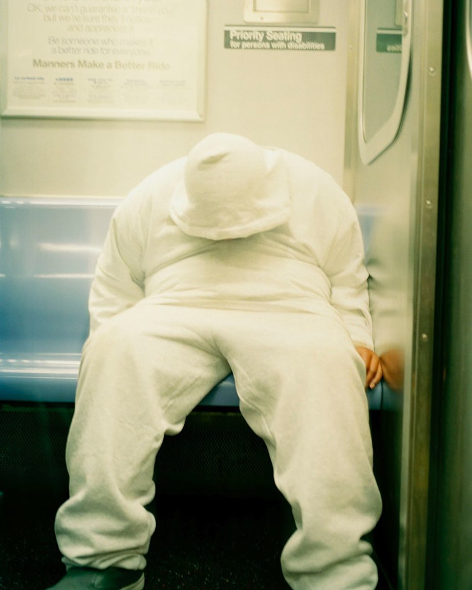 Person slumped on subway seat, wearing hooded sweatsuit, head down, possibly sleeping in an urban transit environment with pale lighting.